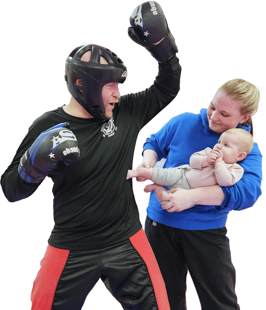 Master Sarah Buzby holds her daughter in position to kick daddy, Master Michael Buzby, who is dressed in sparring gear.